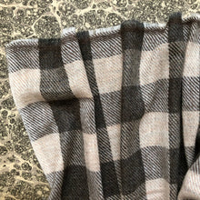 Load image into Gallery viewer, A close-up of a soft woven plaid scarf in gray, brown and warm off-white, draped to show its texture and pattern against a patterned surface.